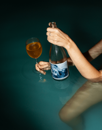 Osoba sedící ve vodě drží láhev vína Alma Wines Anna Serra s modrou etiketou a sklenici s oranžovým vínem.