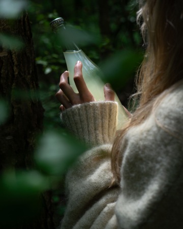 Osoba v béžovém svetru drží láhev březové mízy Euforie uprostřed lesa, jemné světlo proniká skrze listí.