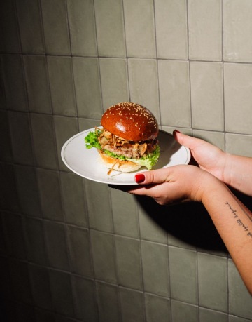 Šťavnatý burger s listem salátu, karamelizovanou cibulkou a sezamovou houskou, servírovaný na bílém talíři v restauraci Dish.