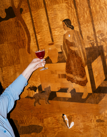 Ruka držící sklenku s červeným koktejlem od Vikeza gastro v rámci Era pop-upu před dřevěnou stěnou s vyobrazením ženy a psa.